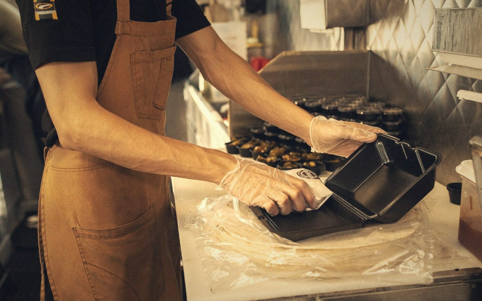Worker in a tan apron packs a takeout container behind a kitchen counter