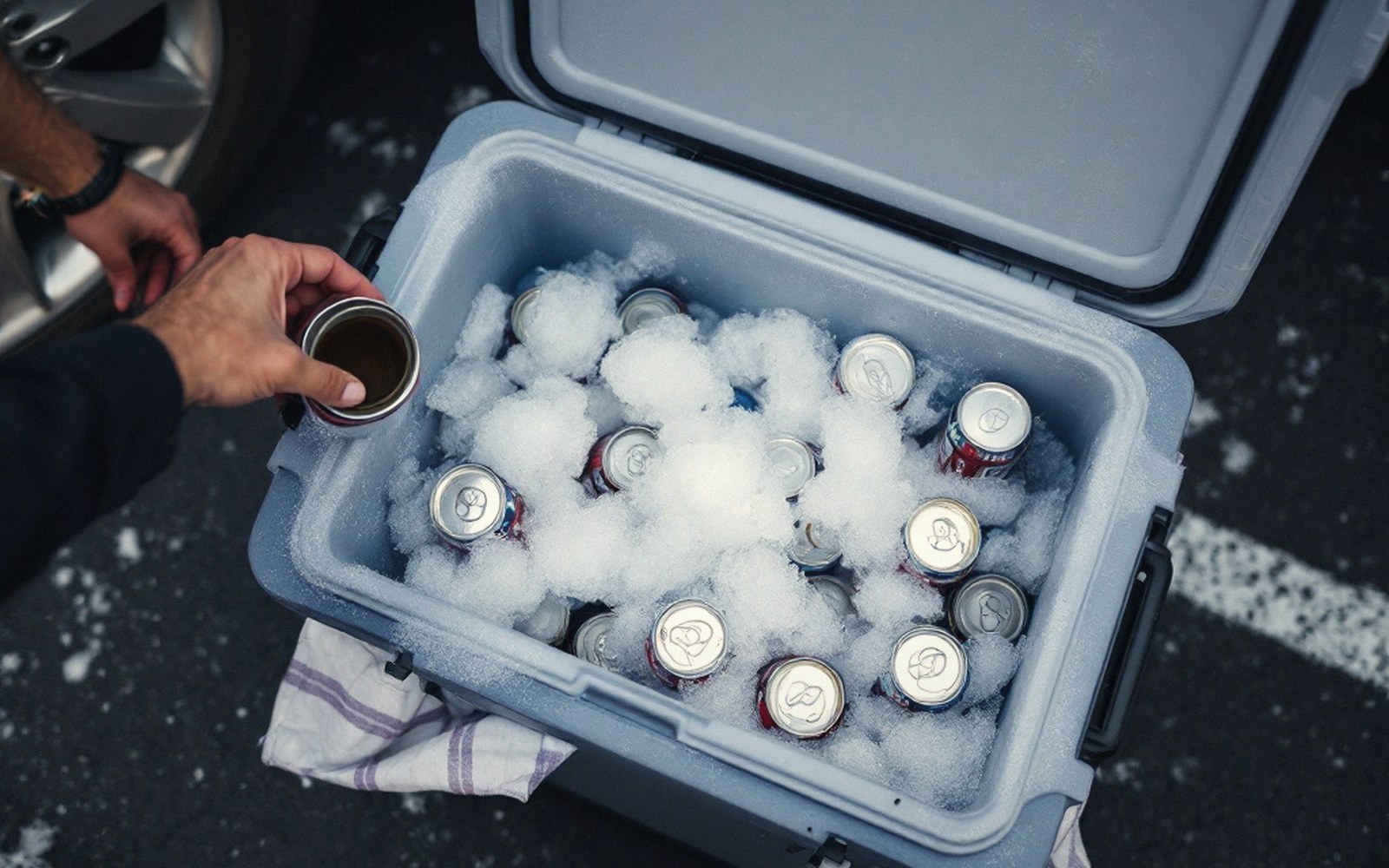 A hand pulls a canned drink from an ice-filled cooler in a parking lot