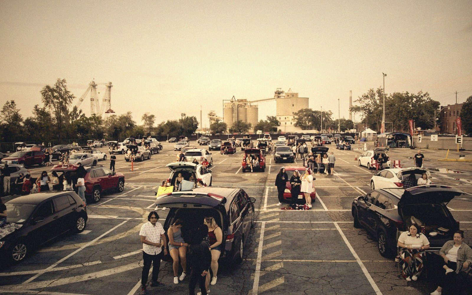 Groups of people tailgate with open car trunks in a parking lot at dusk