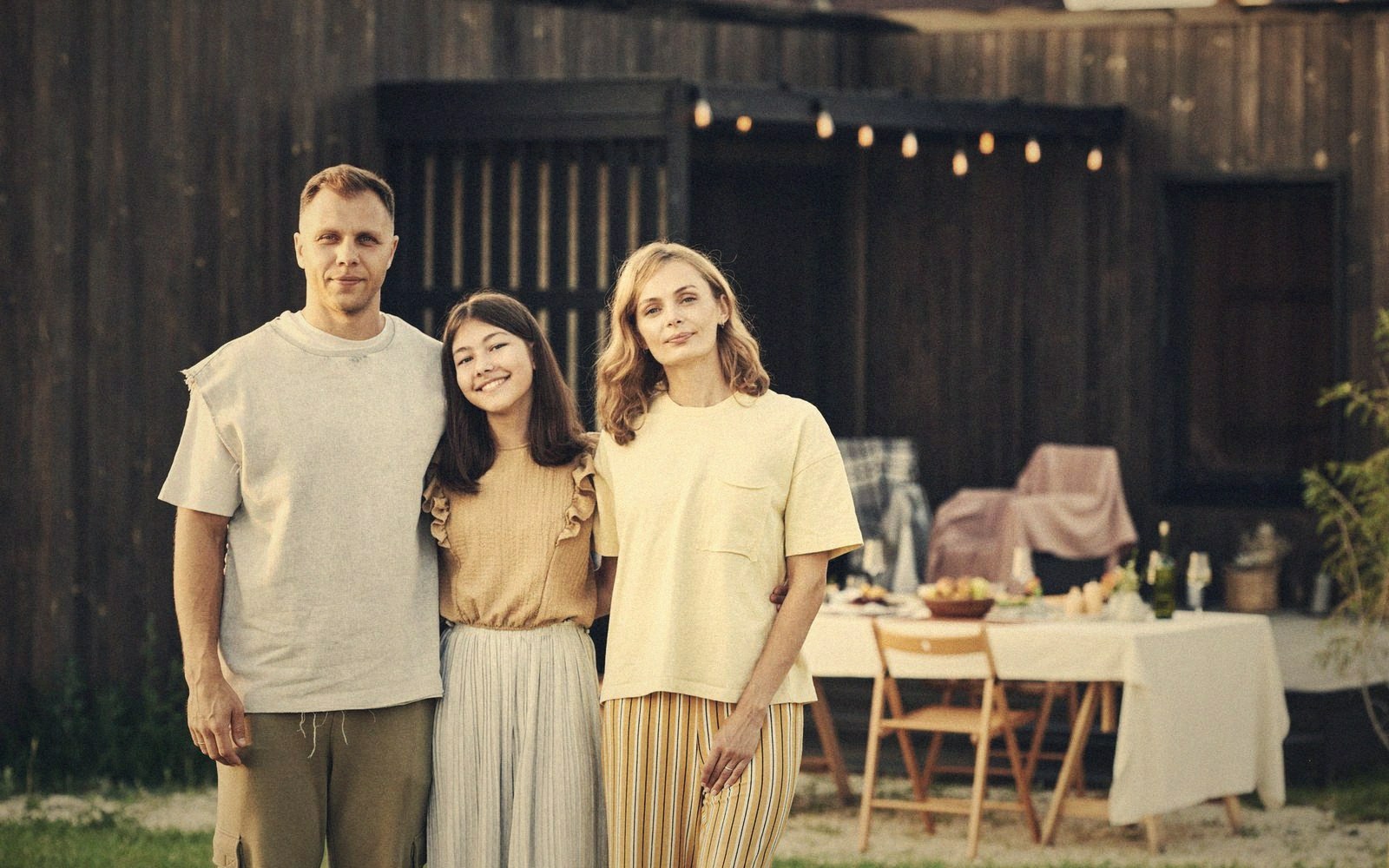 Two adults and a teenager stand together outdoors near a set outdoor dining table