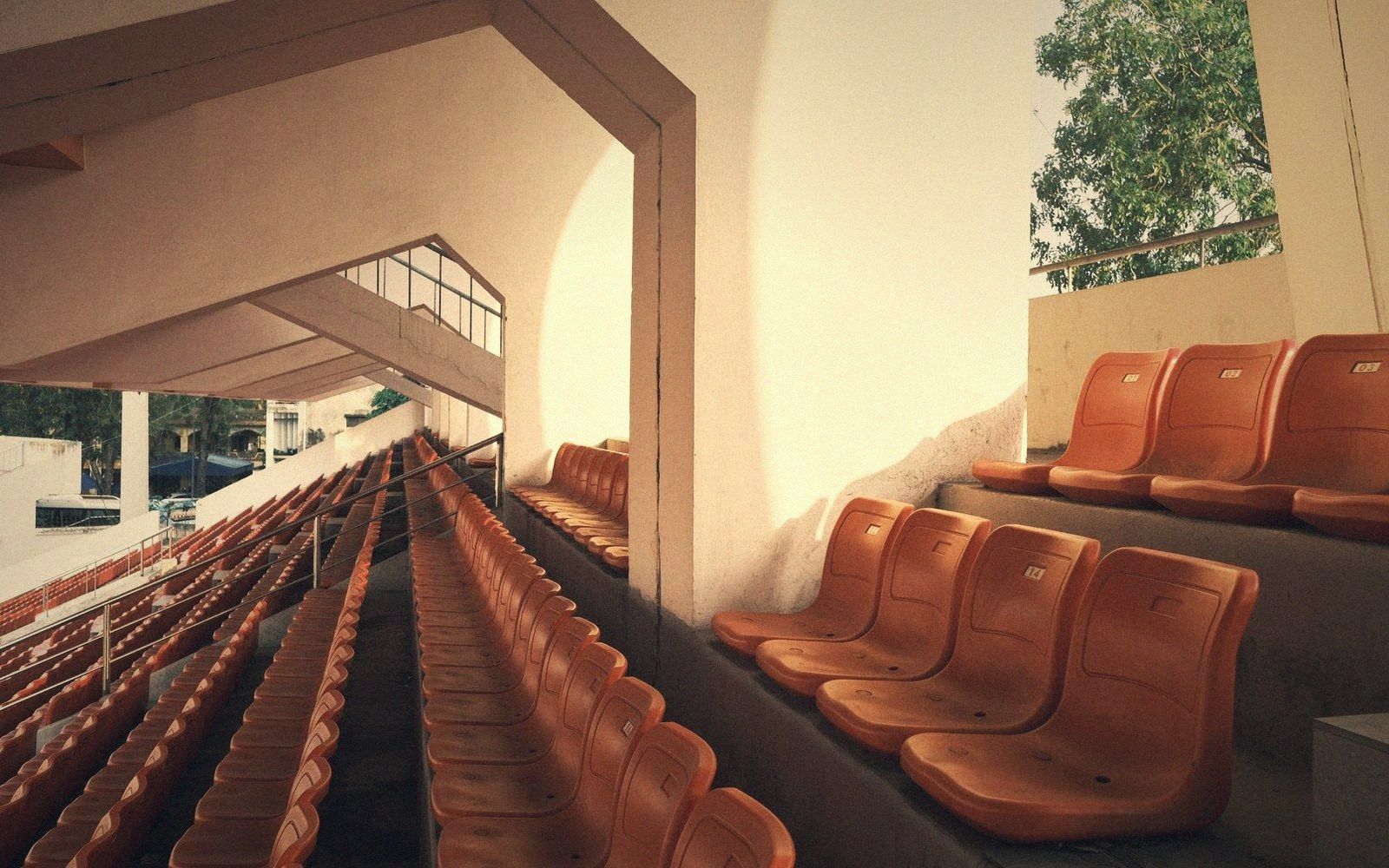 Empty rows of orange plastic stadium seating beneath a covered concrete grandstand